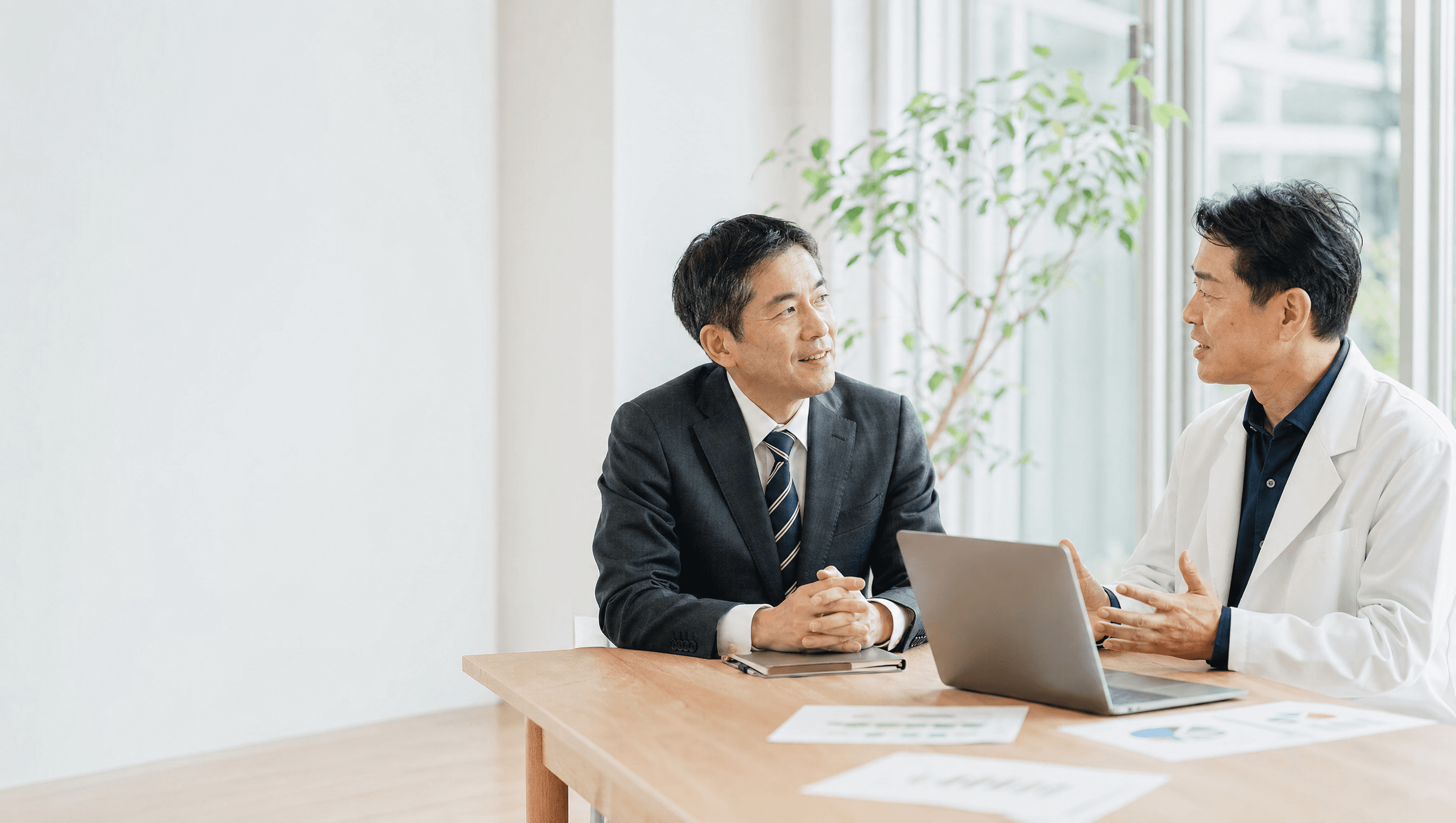 白衣の顧問と経営者が、明るい部屋で同じテーブルに向き合う様子